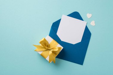 Top view photo of open blue envelope with card small paper hearts and white giftbox with yellow ribbon bow on isolated pastel blue background with copyspace