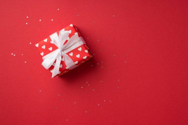 Top view photo of st valentine's day decorations glowing sequins giftbox in red wrapping paper with heart pattern and white ribbon bow on isolated red background with blank space