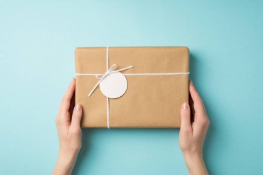 First person top view photo of woman's hands holding craft paper giftbox with small bow and tag on isolated pastel blue background with empty space