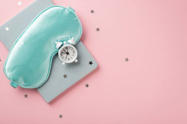Top view photo of blue silk sleeping mask small white alarm clock blue notepad and star shaped confetti on isolated pastel pink background with copyspace