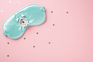 Top view photo of small white alarm clock on blue silk sleeping mask and star shaped confetti on isolated pastel pink background with copyspace