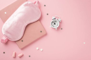 Top view photo of pink silk sleeping mask small white alarm clock pills earplugs pink notebook and star shaped confetti on isolated pastel pink background with copyspace