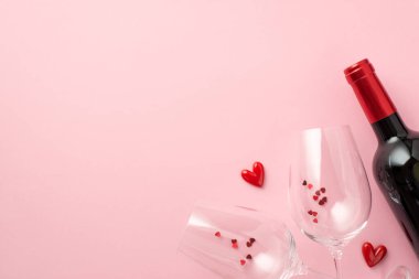 Top view photo of valentine's day decorations small hearts between two wineglasses with confetti and wine bottle on isolated pastel pink background with empty space