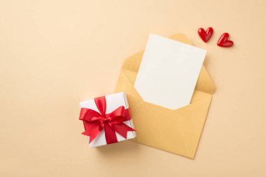 Top view photo of open pastel yellow envelope with paper sheet small red hearts and white giftbox with red ribbon bow on isolated beige background with blank space
