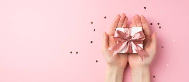 Sevgililer Günü dekorasyonunun birinci şahıs fotoğrafı. Ellerinde pembe kurdeleli küçük beyaz hediye kutusu ve izole edilmiş pastel pembe arka planlı konfeti.
