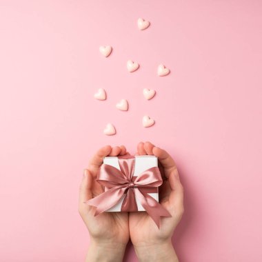 Sevgililer Günü dekorasyonunun dikey fotoğrafında ilk kişi elinde pembe kurdeleli küçük beyaz hediye kutusu ve izole edilmiş pastel arka planda küçük pembe kalpler.