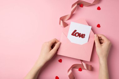 Sevgililer günü dekoratörünün elinde pembe bir zarf tutarken çekilen ilk fotoğrafta kart ve yazılı aşk kırmızı kalpler izole edilmiş pastel pembe arka planda saten kıvrımlı kurdele.