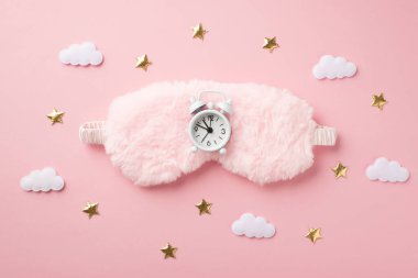 Pembe tüylü uyku maskesinin üzerindeki beyaz alarmlı saatin ve izole edilmiş pastel arka plandaki altın yıldızların en üst görüntüsü.