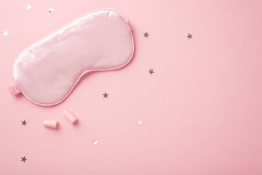 Üst görüntüde pembe ipek uyku maskesi kulak tıkaçları ve izole edilmiş pastel pembe arka planda yıldız şekilli konfeti fotosu var.
