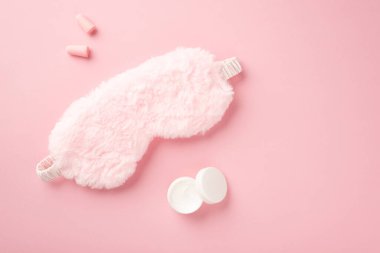 Pembe pofuduk uyku maskesinin üst görüntüsü küçük açık krema şişesi ve kopyalanmış pastel pembe arka planda kulak tıkaçları.
