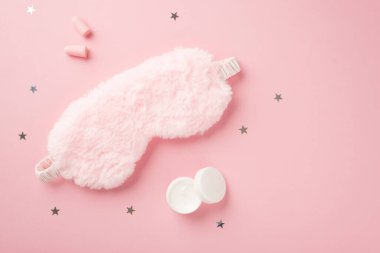 Pembe tüylü uyku maskesinin üst görüntüsü küçük açık krem şişesi kulak tıkaçları ve izole edilmiş pastel pembe arka planda yıldız şeklinde konfeti.