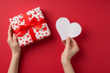 Sevgililer Günü dekorasyonunun birinci şahıs fotoğrafı. Elinde kağıt kalp ve hediye kutusu olan, kalp desenli kırmızı arka planda kırmızı fiyonk ve kopyalanmış kırmızı kaplama olan beyaz ambalaj kağıdı tutan kadın elleri.