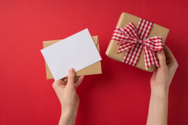 Sevgililer Günü dekoratörünün ellerinin ilk resmi. Zarf ve kareli kurdele ile kapalı kırmızı arka planda boş alan ile el işi kağıt hediye kutusu.