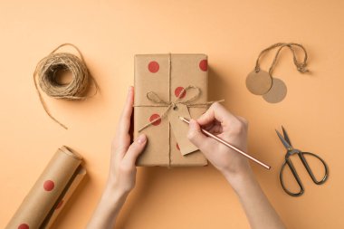 El sanatları araçlarının birinci şahıs fotoğrafı. Kızın elleri etikete iliştirilmiş polka desenli kağıt hediye kutusu ve izole edilmiş bej arka planda sicim yayı.