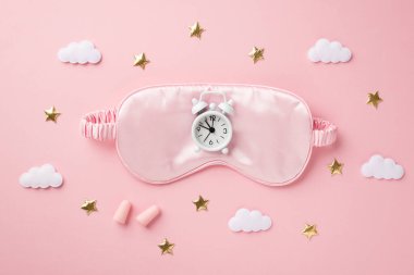 Pembe ipek uyku maskesindeki beyaz alarmlı saatin üst görüntüsü kulak tıkaçları altın yıldızlar ve izole edilmiş pastel arka plandaki bulutlar.