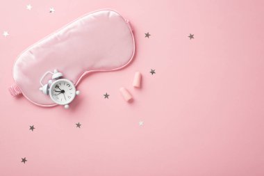 Pembe ipek uyku maskesinin üst görüntüsü küçük beyaz alarmlı kulak tıkaçları ve izole edilmiş pastel pembe arka planda yıldız şeklinde konfeti.