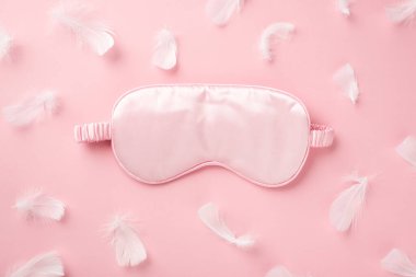 Üst görüntüde pembe ipek uyku maskesi ve açık pembe tüyler, izole edilmiş pastel pembe arka planda, boş alanda.