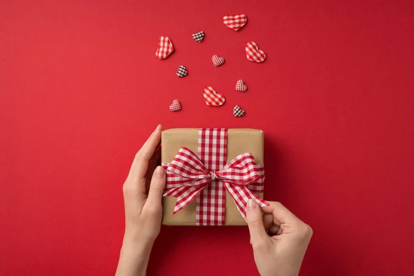 バレンタインデーの装飾の最初の人のトップビュー写真女の子の手チェックリボン弓と孤立した赤の背景に心とクラフト紙のギフトボックスをアンラッピング — ストック写真