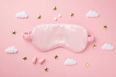 Üst görüntüde pembe ipek uyku maskesi kulak tıkaçları haplar altın yıldızlar ve bulutlar kopyalanmış pastel pembe arka planda