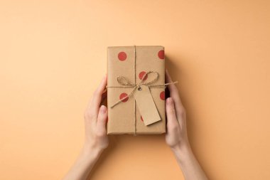 Sevgililer Günü dekorasyonunun birinci şahıs fotoğrafı. El sanatları el sanatları hediye kutusu, polka desenli etiket ve kopyalanmış bej arka planda sicim yayı.