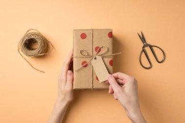 Sevgililer Günü dekorasyonunun birinci şahıs fotoğrafı el işi kağıt hediye kutusuna polka desenli sicim ve makas makasıyla dokunan kadın elleri kopyalanmış bej arka planda.