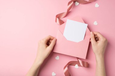 Sevgililer günü dekoratörünün elinde pembe bir zarfla kalp şeklinde konfeti ve izole edilmiş pastel pembe arka planda pembe saten kıvrımlı kurdele tutan ilk kişinin fotoğrafı.