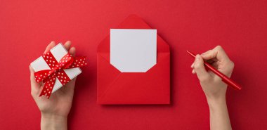 Sevgililer Günü dekorasyonunun birinci şahıs fotoğrafı. Küçük hediye kutusu ve kalem kağıdın üzerinde boş bir kırmızı zemin ve izole edilmiş kırmızı bir zarfın içinde.