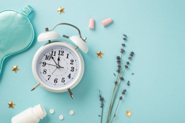 Beyaz alarm saatinin üst görüntüsü lavanta mavisi saten uyku maskesi pembe kulak tıkaçları haplar ve izole edilmiş pastel mavi arka planda altın yıldızlar.
