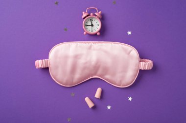 Büyük ipek uyku maskesinin üst görüntüsü ve üzerinde dağınık yıldız şeklinde konfeti ve menekşe arka planında iki pembe kulak tıkacı olan küçük pembe saat.