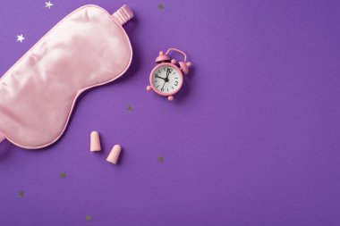 Büyük ipek göz maskesi ve mor izole arka plandaki yıldız şeklinde konfetileri olan iki kulak tıkaçlı sevimli pembe saatin en üstteki fotoğrafı.