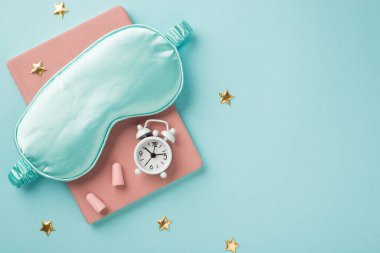 Üzerinde beyaz alarm saati ve kulak tıkaçları olan pembe kitaptaki büyük ipek göz maskesinin üst görünümü altın yıldızlı ve kopyalanmış mavi arka planda.