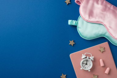 Üzerinde pembe ve açık mavi iki ipek maske, bir pembe günlük ve beyaz alarm ve iki kulak tıkacı ve derin mavi izole arka planda parlak yıldız şeklinde bir konfeti olan boş bir alan.