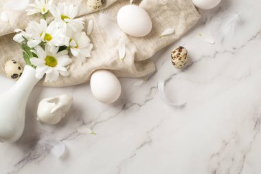 Üzerinde papatya yaprakları ve tüyler serpiştirilmiş vazonun, izole edilmiş mermer arka planda desenli kumaşın üzerinde birkaç yumurta ve seramik tavşan resmi.