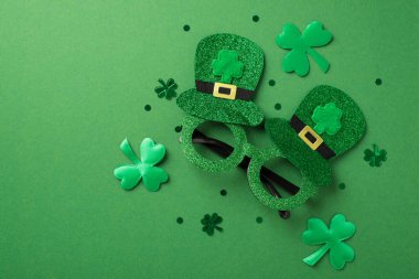 Yeşil, abartılı, parlak gözlüklü, üzerinde iki şapka olan fotografın ve yeşil izole arka planda farklı boyutlarda yonca şekilli konfetilerin üst görüntüsü.