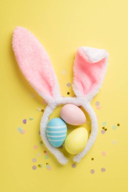Üç pastel renkli Paskalya yumurtası ve izole edilmiş sarı arka planda şirin konfetileri olan tavşan kulağı şeklinde yumuşak saç bandı.