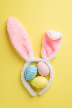 Üç pastel renkli Paskalya yumurtası ve izole edilmiş sarı arka planda tavşan kulağı şeklinde şirin bir saç bandı.