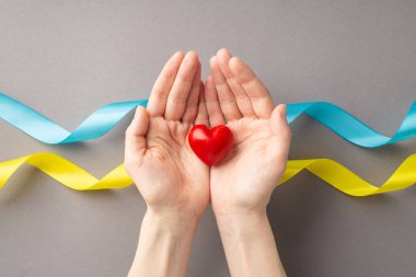 Ukrayna 'daki savaşı durdurun. Kadın ellerinin avuçlarında küçük kırmızı bir kalple ulusal bayrak renklerinde gri arka planda kıvrımlı kurdeleler taşıdığı üst tepedeki görüntü.