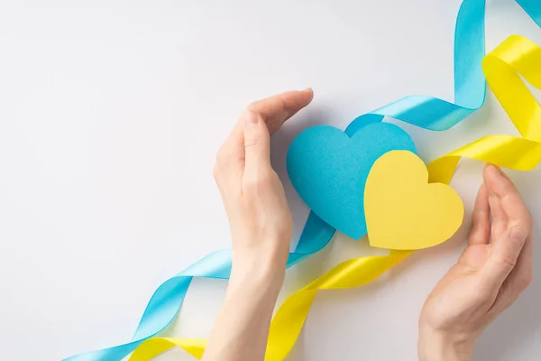 Stop the war in Ukraine concept. First person top view photo of young woman\'s hands protecting two yellow and blue hearts and wavy ribbons on white background