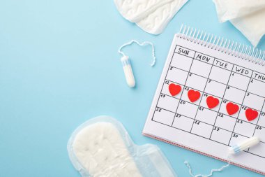 Takvimdeki kırmızı kalp işaretlerinin üst görüntüsü hijyenik peçeteler ve tamponlar izole edilmiş pastel mavi arka planda kopyalanmış