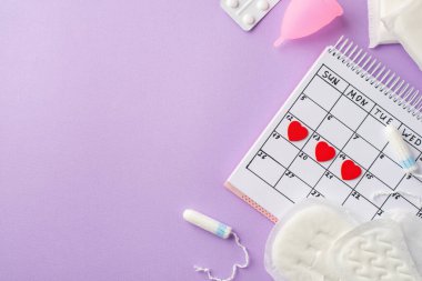 Takvimdeki kırmızı kalp izlerinin üst görüntüsü pembe regl bardağı tabağı hijyenik peçeteler ve izole edilmiş pastel menekşe arkaplanlı tampon.