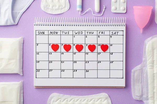 Takvimdeki kırmızı kalp izlerinin üst görüntüsü iç çamaşırı pembe adet bardağı ağrı kesici hijyenik pedler ve izole edilmiş pastel leylak arka planda tamponlar.