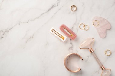 Cilt güzellik konsepti. Pembe saç tokalarının üst görüntüsü gül kuvars roller gua sha altın halkalar ve beyaz mermer arka planda bilezik kopyalanmış