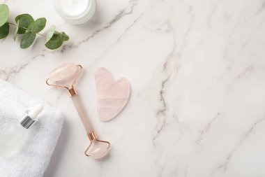 Cilt bakımı. Gül kuvars roller gua sha masajcı krem kavanozu şeffaf şişe okaliptüs ve beyaz havlunun beyaz mermer zemin üzerinde telif alanı