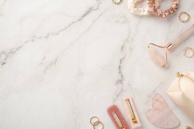 Cilt güzellik konsepti. Beyaz mermer arka planda pembe tokaların, gül kuvars roller gua sha altın yüzüklerin, saç tokalarının ve kozmetik çantalarının resmi.