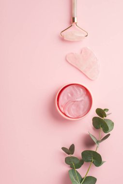 Güzellik prosedürü konsepti. Rose Quartz roller gua pembe göz bantları ve okaliptüs filizi dikey görüntüsü izole edilmiş pastel pembe arka plan