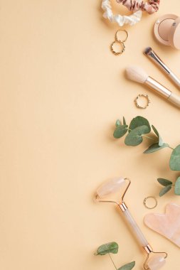 Altın yüzüklerin dikey fotoğrafı göz makyajı fırçaları gül kuvars silindiri gua sha tokaları ve izole edilmiş pastel bej arka plan üzerinde okaliptüs.