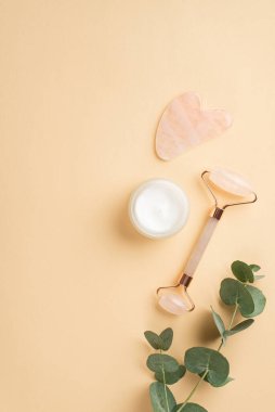 Güzellik prosedürü konsepti. Rose Quartz roller gua sha masajcı cam krem kavanozu ve izole edilmiş pastel bej arka planda okaliptüs dalının dikey görüntüsü