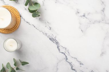 Top view photo of candles on rattan serving mat and eucalyptus on white marble background with copyspace