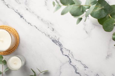 Top view photo of candles on rattan serving mat and ceramic vase with eucalyptus on white marble background with copyspace