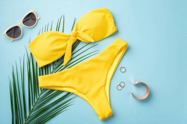 Yaz tatili kavramı. Altın yüzüklerin üst görüntüsü bilezik stili güneş gözlüğü sarı bikini ve izole edilmiş pastel mavi arka planda palmiye yaprakları.